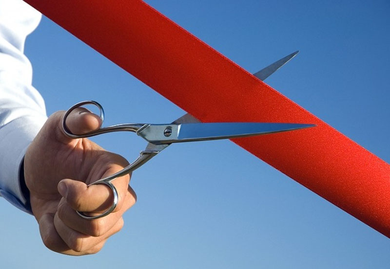 Imagem de uma pessoa segurando uma tesoura e cortando uma faixa vermelha sob um céu azul.